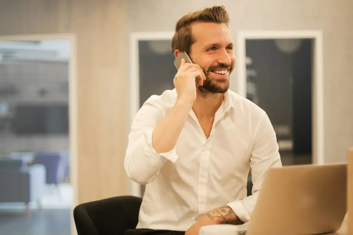 01482 Hull Phone Numbers Smiling bearded man in a white shirt on a mobile phone call at a desk with a laptop and a coffee cup