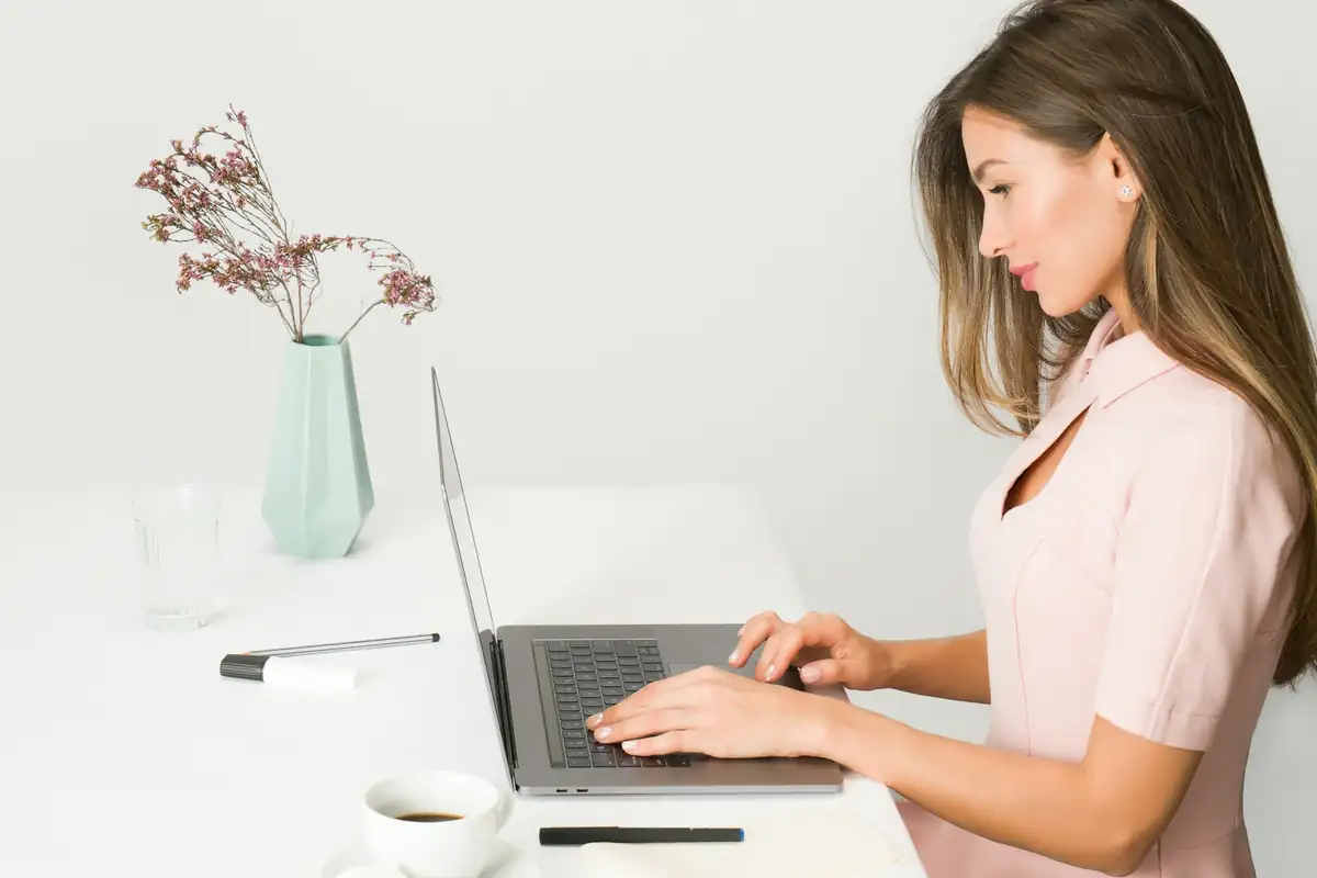 01246 Chesterfield Phone Numbers Woman in a pink top typing on a laptop at a white desk beside a small vase of dried flowers